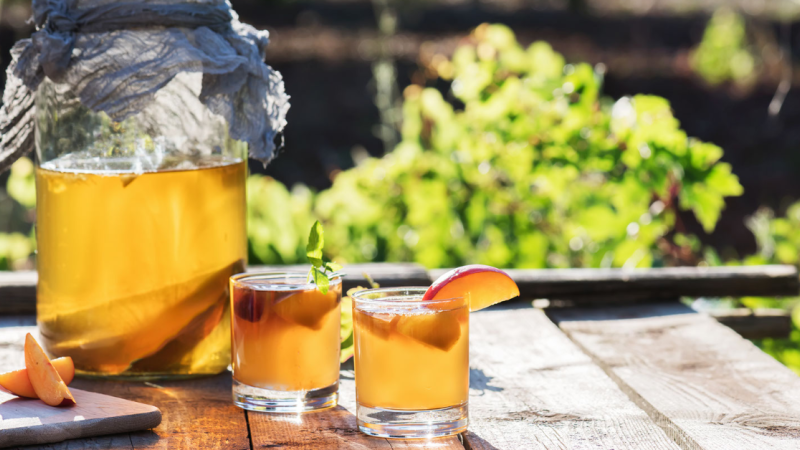 A jar of homemade kombucha and two glasses filled with it sit on an outdoor table.