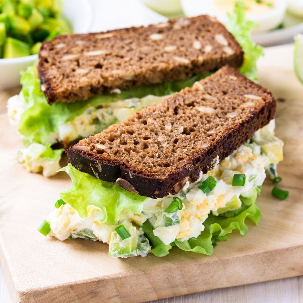 An egg salad sandwich on whole wheat bread sits on a cutting board waiting to be served. Recipes with pickled eggs call for swapping out hard-boiled eggs in this classic sandwich. 