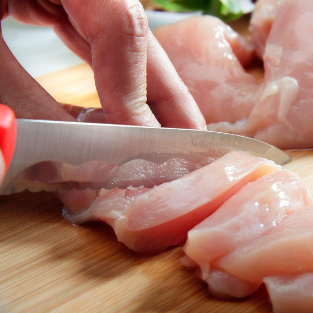 To can raw chicken, you have to cut it up into 1-inch cubes like this woman is doing. She's using  a sharp knife and a sterilized cutting board to cut the raw chicken. 