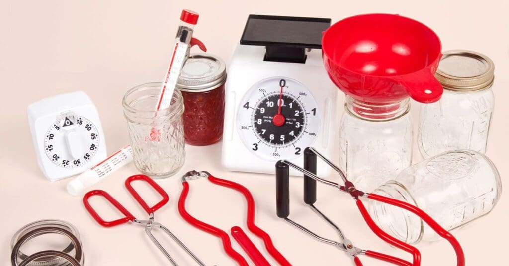 Canning tools are situated on a table to can raw chicken. They include a timer, a thermometer, canning jars with lids, a measuring cup, a funnel, and different size tongs.