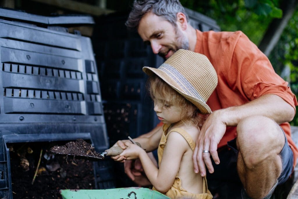 A man and his young daughter use an access door in the side of a compost bin to harvest finished compost. Learning how to compost includes choosing a bin with easy access. 